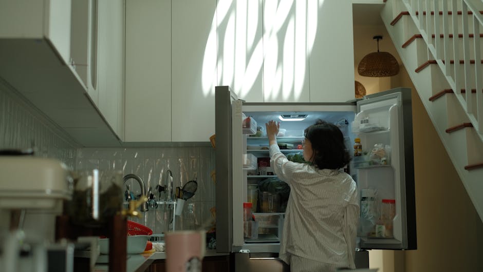 A woman with dark hair wearing a light-colored, long-sleeved shirt is seen from behind as she reaches into a modern, open refrigerator in a domestic kitchen. The fridge is situated against a wall beneath tall, white kitchen cabinets that extend to the ceiling. The door of the fridge is open, revealing various food items, bottles, and containers organized inside on shelves and door compartments. Light from a nearby window casts shadows across the kitchen, creating a pattern on the upper cabinets. To the right of the fridge, a staircase with wooden steps and white risers is visible, leading upwards. On the kitchen countertop, blurred objects such as a pink mug, a cutting board, and small appliances are partially visible, indicating a typical home environment. The context suggests a scene related to packing or preparing for a home relocation, fitting with professional removals services by Man with Van Kenton, as depicted in the image.