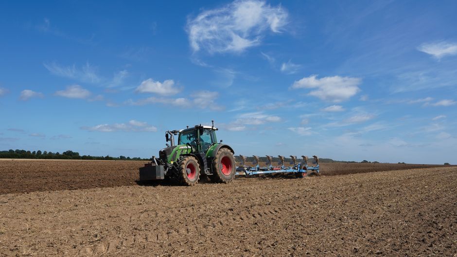 A large green tractor with red wheels is operating on a wide, open farmland under a bright blue sky with scattered white clouds. The tractor is attached to farming equipment used for soil cultivation or planting, positioned on a freshly ploughed section of the field. The scene is outdoors, with the horizon line visible in the distance, and no other machinery or structures present. The image highlights rural agricultural activities related to land preparation for planting or harvesting, demonstrating typical farming work in a spacious, open environment. This setting reflects the scale and nature of vehicle use in agricultural land management, consistent with transportation and logistical considerations in rural property or farmland transitions, relevant to moving or site preparations documented by Man with Van Kenton.