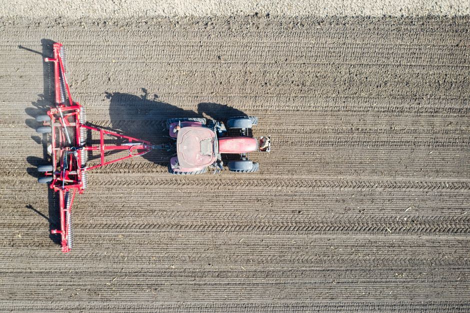 An aerial view of a large tractor fitted with a red agricultural sprayer attachment, positioned on a freshly tilled, expansive field with visible parallel furrows. The tractor, painted in red and gray, is centered in the image, with the sprayer extending to the left and occupying a significant portion of the scene. The field appears in early stages of cultivation, with loose soil and evenly spaced lines, suggesting preparation for planting. Bright sunlight illuminates the scene, casting shadows from the tractor and sprayer onto the soil. This image captures the process of land preparation in rural farming, relevant to the context of house removals and relocation services by Man with Van Kenton, highlighting practical transportation and handling of equipment during moving and setup scenarios.