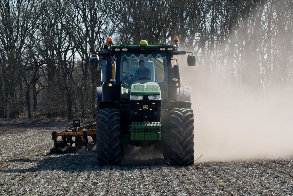A large green tractor with red wheels is operating on a wide, open farmland under a bright blue sky with scattered white clouds. The tractor is attached to farming equipment used for soil cultivation or planting, positioned on a freshly ploughed section of the field. The scene is outdoors, with the horizon line visible in the distance, and no other machinery or structures present. The image highlights rural agricultural activities related to land preparation for planting or harvesting, demonstrating typical farming work in a spacious, open environment. This setting reflects the scale and nature of vehicle use in agricultural land management, consistent with transportation and logistical considerations in rural property or farmland transitions, relevant to moving or site preparations documented by Man with Van Kenton.