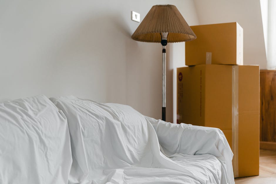 Interior view of a room during a home relocation process, featuring three cardboard moving boxes stacked near a wooden cabinet and a standing floor lamp with a pleated lampshade. The boxes are sealed with packing tape and sit on the wooden floor, with one positioned on top of another. A white sofa covered with a protective fabric is partially visible in the foreground, indicating furniture being prepared for transport. The scene is illuminated by natural light coming through a nearby window, with a plain light-colored wall in the background. This setup reflects the packing and loading process involved in house removals, as carried out by companies like Man with Van Kenton, focusing on organizing and safeguarding belongings before their furniture transport to a new home.