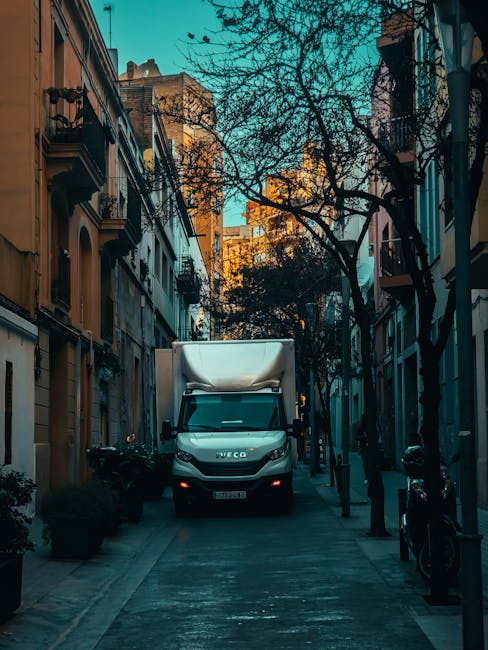 A white Iveco moving van is parked on a narrow urban street between multi-storey buildings with decorative balconies and window frames. The vehicle's cargo area is covered with a white tarp, indicating it is in the process of home relocation or furniture transport. The street is lined with leafless trees, some of which have orange and yellow foliage visible against a clear blue sky. On the pavement, several black bollards and parked motorcycles are present, with a few potted plants near the building entrances. The scene suggests a typical city move involving careful loading or unloading of boxes, furniture, and moving equipment, reflecting the logistical aspects of household removals carried out by companies like Man with Van Kenton. The ambient lighting indicates daylight hours, and the overall setting emphasizes the urban environment suited for professional packing and moving services.