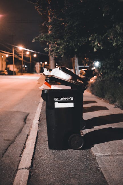 A black wheeled bin labeled 'ST. JOHN'S' positioned on the pavement beside a residential street at night, with a partially open lid containing cardboard and plastic packaging materials. The scene features street lighting casting a warm glow, with trees and bushes lining the sidewalk, and distant houses faintly visible. The bin appears to be used for waste disposal during a home relocation or moving process, reflecting typical rubbish collection practices in Kenton. This image illustrates the importance of clearing waste before a house removal with professional services such as those provided by Man with Van Kenton, focusing on proper disposal during the moving process and adherence to local rubbish rules outlined in the Kenton Council guidelines for rubbish disposal during moves.