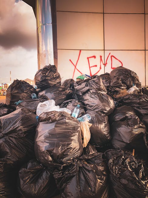 A large pile of black garbage bags filled with waste, stacked against a tiled wall on a pavement. Some bags are torn or bulging, revealing plastic bottles and other rubbish inside. The bags are located outdoors during daylight, with a partly cloudy sky visible in the background. Red spray-painted graffiti saying 'XEND' is visible on the wall behind the waste. This scene illustrates improper rubbish disposal, which can occur during house moves when waste is accumulated or discarded incorrectly. Man with Van Kenton offers expert removals and packing services to ensure proper waste management during relocations, aligning with local rubbish rules and environmental guidelines.