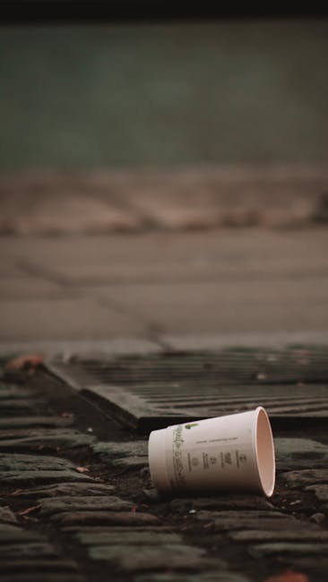 A black wheeled bin labeled 'ST. JOHN'S' positioned on the pavement beside a residential street at night, with a partially open lid containing cardboard and plastic packaging materials. The scene features street lighting casting a warm glow, with trees and bushes lining the sidewalk, and distant houses faintly visible. The bin appears to be used for waste disposal during a home relocation or moving process, reflecting typical rubbish collection practices in Kenton. This image illustrates the importance of clearing waste before a house removal with professional services such as those provided by Man with Van Kenton, focusing on proper disposal during the moving process and adherence to local rubbish rules outlined in the Kenton Council guidelines for rubbish disposal during moves.