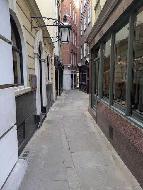 A narrow urban alleyway with a paved stone surface, flanked by multi-storey buildings on either side. On the left, there are cream and grey coloured building facades with arched windows and small stone ledges, while on the right, a shopfront with large glass windows displaying clothing and accessories. The alley appears to be during daytime with diffused natural light. At the far end, the alley curves slightly out of view, showing a mixture of brick and stone exterior facades. There are no visible people or moving equipment, but the scene reflects a typical location for home relocation or furniture transport services such as those provided by Man with Van Kenton, especially in context of tight residential streets related to the Kenton HA3 area. This setting is indicative of a typical UK residential street suitable for house removals and packing and moving activities.