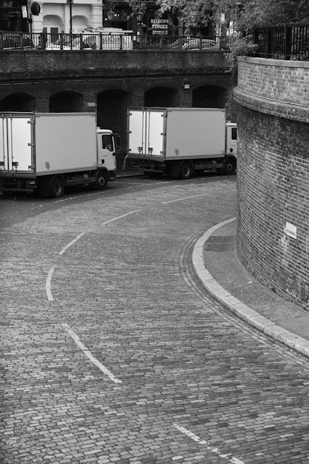 A black and white photograph of a narrow, curved cobblestone street in an urban area, with two large moving vans parked along the roadside. The vans are adjacent to a brick wall with a small sign, and their rear doors are visible, indicating they are prepared for loading or unloading. Inside the vans, cardboard boxes, plastic-wrapped furniture, and packing materials can be seen through the open rear doors, suggesting a home relocation or furniture transport process. The street is lined by a terrace with railings and trees, and there is an underpass or bridge with arched openings above the vans, highlighting a multi-level environment typical of city logistics. This scene reflects the practical aspects of house removals and moving services, such as vehicle preparation and maneuvering through narrow lanes, as managed by companies like Man with Van Kenton.