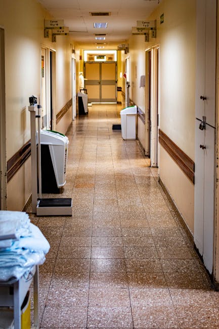 Inside a corridor of a residential or healthcare facility, with beige tiled flooring and cream-colored walls, featuring wooden skirting boards. On the left side, there is a white and gray portable air