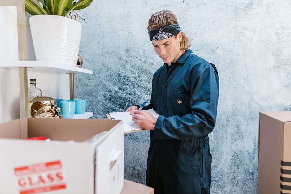 A young man dressed in a dark blue uniform with a black and white bandana on his head is inside a room, holding a clipboard and pen while inspecting or taking notes during a home relocation process. Behind him, there are white shelves with decorative items such as a large white ceramic pot with a green plant, a glass decanter, and blue ceramic cups. In the foreground, a cardboard box with 'Class' printed on it is partially visible, containing various items. To the right, there are additional cardboard boxes stacked on the floor, indicating an ongoing packing or unpacking phase. The room has a textured light blue wall and is lit with natural light, suggesting a daytime move. This scene reflects the packing and moving process typical of professional removals services, as seen in Man with Van Kenton, highlighting the careful handling and organization involved in home furniture transport and packing for a house move, especially when relocating out of Kenton Road.