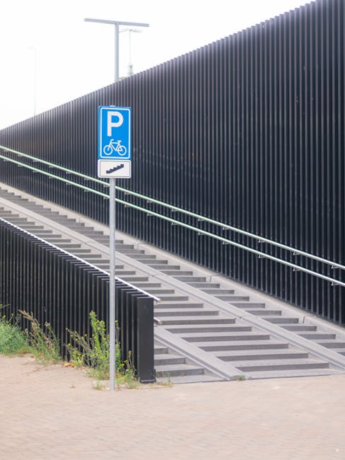 A sloped outdoor access ramp made of concrete with metal railings on both sides, designed for wheelchair and pedestrian use, positioned adjacent to a dark-colored fencing panel. The ramp features even