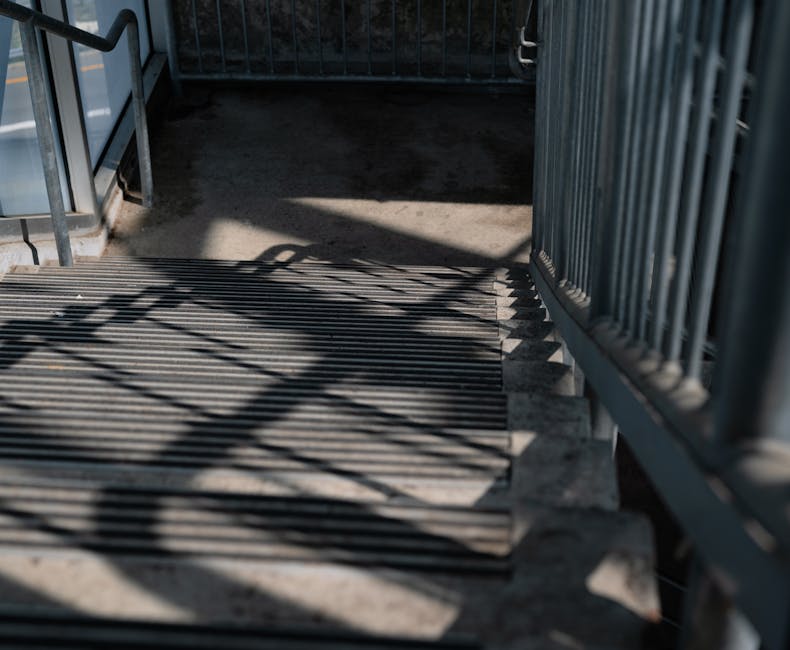 A sloped outdoor access ramp made of concrete with metal railings on both sides, designed for wheelchair and pedestrian use, positioned adjacent to a dark-colored fencing panel. The ramp features even