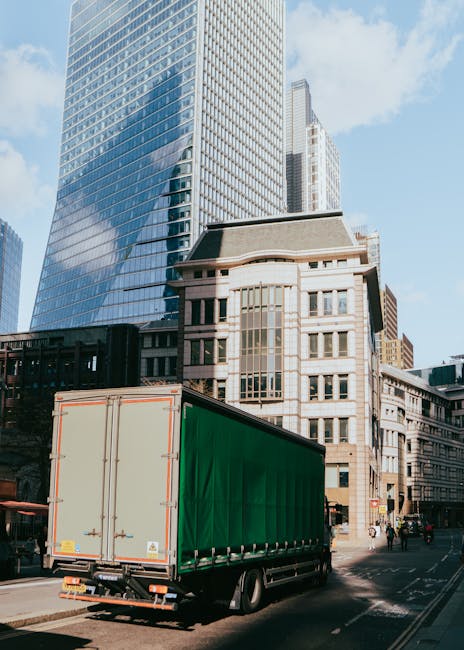 A large moving truck with a green cargo area and beige rear doors is parked on an urban street, positioned close to the sidewalk with a loading area for home relocation services. The truck is situated
