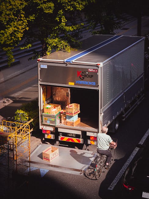 A large metallic moving truck with a partially open rear door, parked on a street during daylight hours, is being loaded with cardboard boxes and plastic crates containing household items. The boxes are of various sizes, some taped and others without, with packing materials visible around them. A man wearing casual clothing and a helmet is riding a bicycle beside the truck, possibly assisting with the move. Nearby, a yellow metal cage or trolley is positioned on the sidewalk, awaiting use in the home relocation process. The truck is situated next to a curb, with trees and a building entrance visible in the background, indicating an urban environment suitable for a professional removals service such as Man with Van Kenton. This scene depicts the loading process of furniture transport and packing during a move, highlighting the logistics involved in house removals and last-minute booking options.