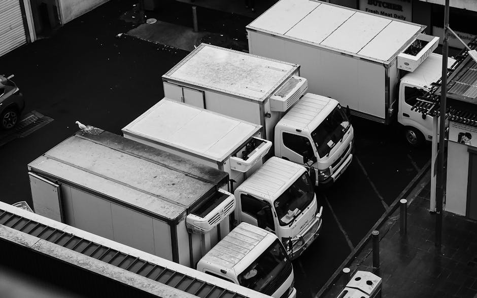 Aerial view of several white delivery vans and box trucks parked closely together in a loading area outside a commercial building. The vehicles are arranged in parallel, with some showing visible roof racks and cargo doors, indicating they are used for home relocation or furniture transport. The surrounding environment includes a paved surface, bollards, and part of an adjacent structure with a sign and store entrance, suggesting an urban setting. The trucks are positioned to facilitate loading and unloading processes, with no visible personnel or active moving actions. The image highlights the logistical aspect of packing and moving services, such as those provided by Man with Van Kenton, emphasizing the use of multiple vehicles for a house relocation or furniture transport task.