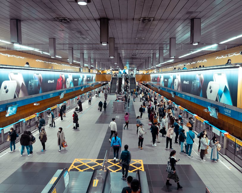 A busy indoor train station with a high ceiling and bright lighting, featuring a wide central walkway filled with numerous people, some walking, others waiting or using smartphone devices. On the upper level, there are travel advertisements displayed on large electronic billboards featuring images of smartphones and gadgets. The lower floor has signs and ticketing machines along the sides, with a set of escalators and stairs in the middle connecting the two levels. The environment appears modern and spacious, indicating a transportation hub where home relocation or moving services like those offered by Man with Van Kenton could be relevant for assisting with the transfer of furniture or belongings during a move near Kenton station.
