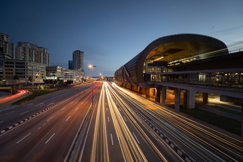 A long-exposure photograph capturing a multi-lane urban road at dusk, with streaks of white and red light trails created by moving vehicles. To the right, there is a modern, curved station building with large glass windows and a covered platform, elevated above the road and supported by concrete pillars. In the background, tall residential and commercial buildings line the skyline, illuminated by internal and external lights. The scene is lit by streetlights and the fading natural light of the early evening, depicting an active city environment. This image reflects aspects of urban infrastructure relevant to home relocation and moving services, such as transportation and logistics, as seen through the movement and architectural context, which are areas covered under [PAGE_TITLE] about moving near Kenton Station. Man with Van Kenton occasionally assists with furniture transport and packing during such urban relocations.